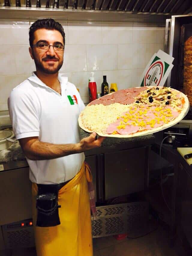 Herr Ekrem Tokat hält eine Pizza in beiden Händen die gleich in den Pizzaofen kommt.